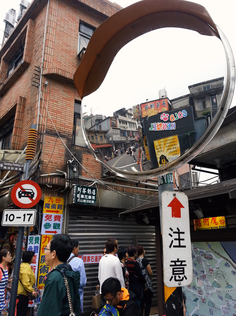 A photo of a street scene with people, a convex mirror, a sign with the word 'Jiufen', a directional sign with '出口', and a map with a 'Jiufen' label.